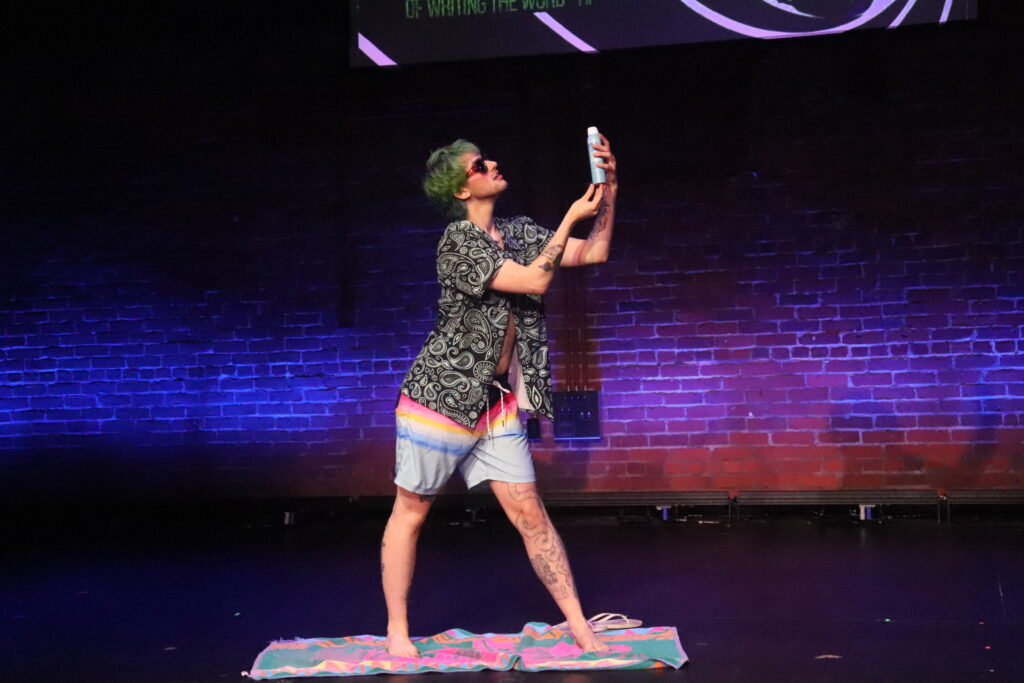 Drag performer James Bondage creates a beach scene on stage, presenting spray-on sunscreen to the audience. He is wearing sunglasses and swim trunks.