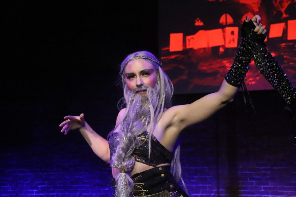 Providence-based burlesque performer Anita Tension removes a black glove during her performance. She has a long gray wizard wig and beard and is wearing a black bandeau top.