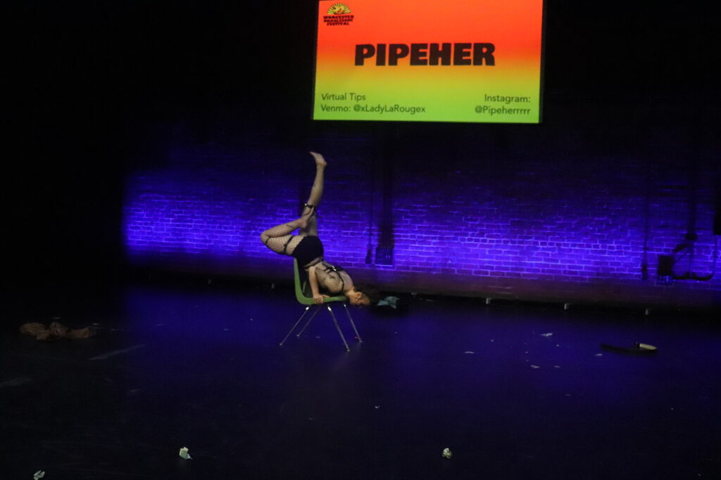 Pipeher balances over a chair on stage, legs in the air over their back. They are wearing a fishnet bodysuit and harness.