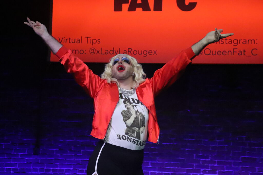 Worcester-based drag queen Fat C poses on stage with her arms spread open. She has bold red lipstick and blue eyeshadow, stubble, and blond wig, wearing a red jacket and Linda Ronstadt T-shirt