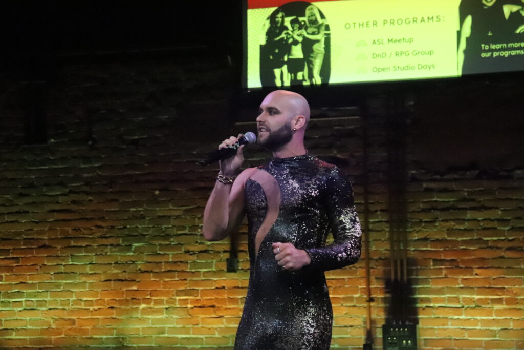 Love Your Labels Executive Director Joshua Croke speaks from the stage during intermission. They are bald with a short beard, wearing a black sequin gown with cutouts.
