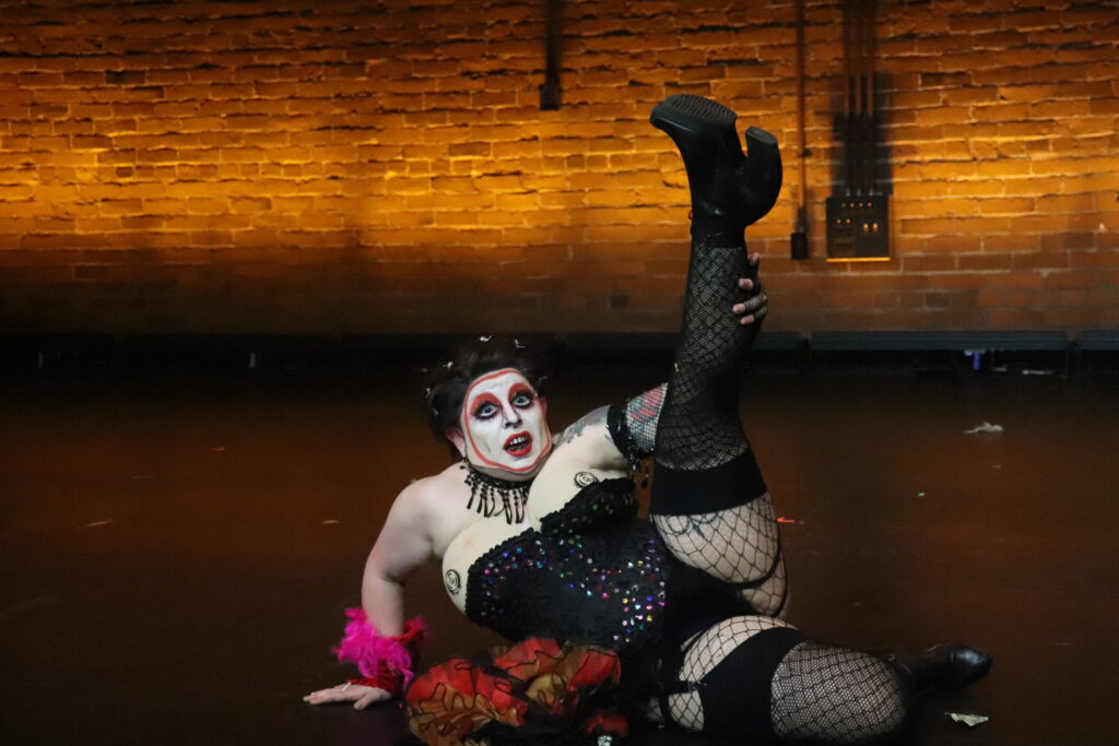 Boston-based drag performer SHERMAN stretches their leg in the air on stage. They are wearing black fishnets, chunky heels, a corset, and red boa.