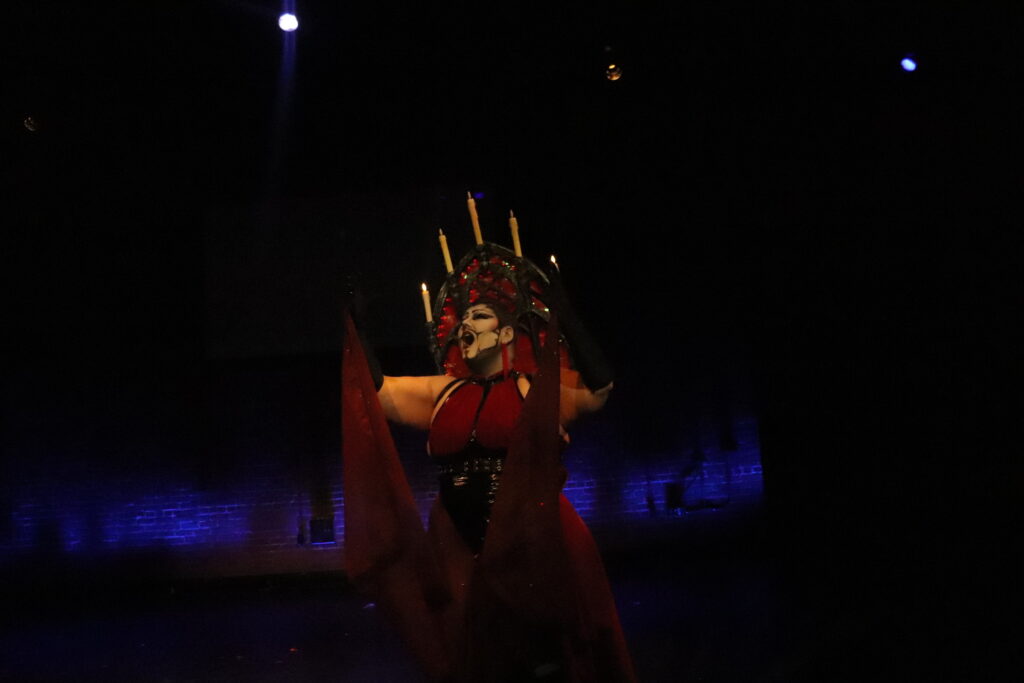 Drag performer Darren TaShine lipsyncs opera on stage. They have eerie white and gray makeup, and wearing a headpiece that looks like a gothic stained-glass window and candelabra.