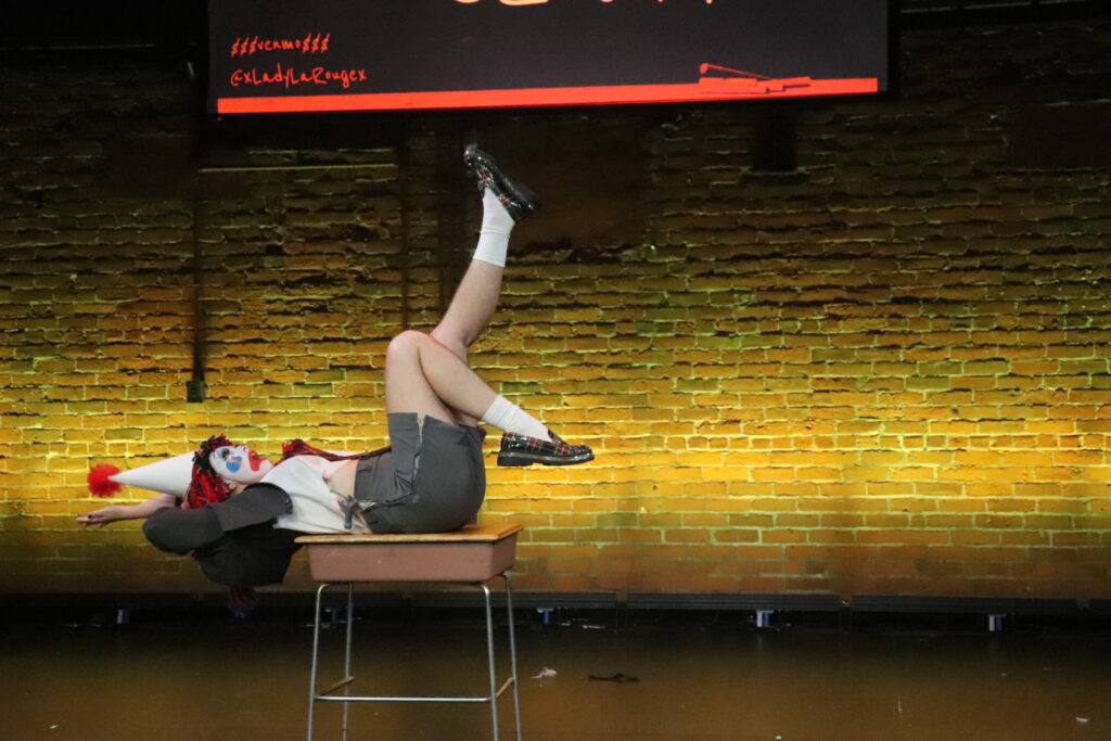 Cheerful the Clown is laying on a school desk, kicking their feet in the air. They are wearing a school uniform with shorts and a blazer, and a white dunce cap on their red wig made from yarn.