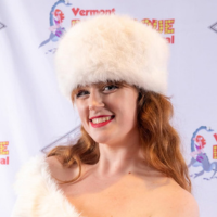 Caucasian woman with red hair smiling and wearing a fuzzy white winter hat in front of a step and repeat